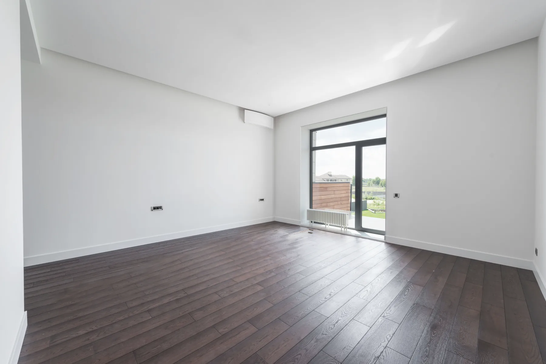 Großer leerer Wohnraum mit dunklem Holzboden und Tür zu einer Terrasse